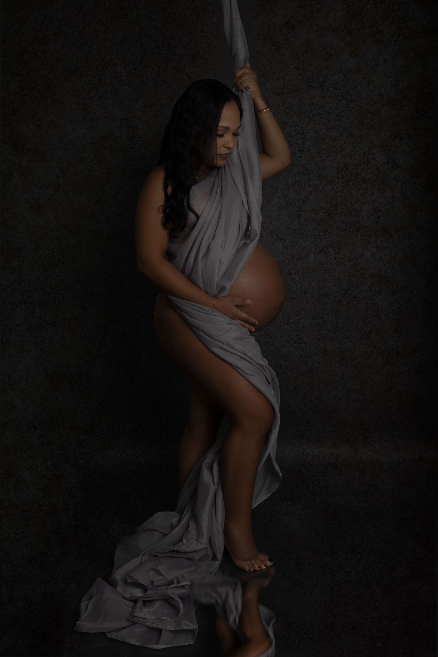 Pregnant woman draped in flowing gray fabric poses gracefully in a dark studio maternity photography session.