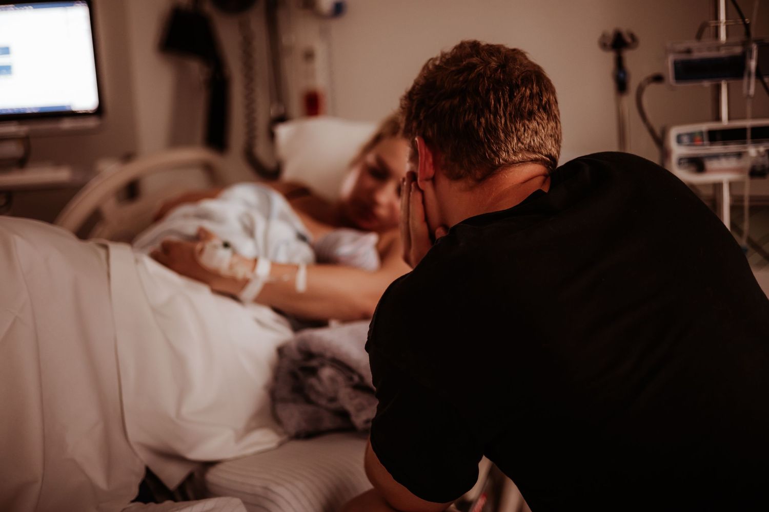 Emotional partner kneeling beside hospital bed, hands clasped in prayer while supporting laboring woman during childbirth.