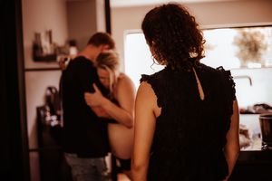 A woman in black watches a couple embracing in a warmly lit kitchen during a birth doula support session.
