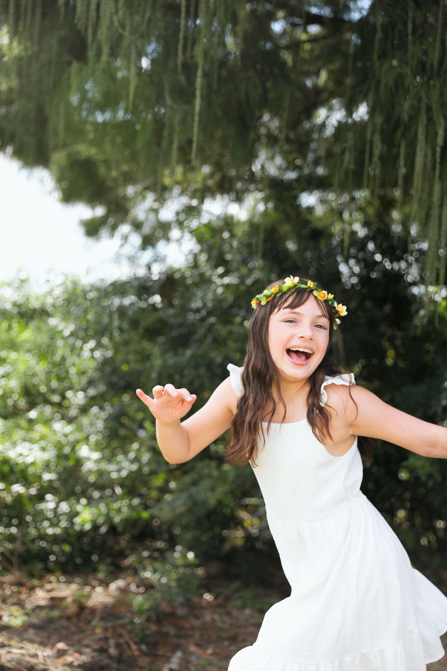Joyful girl in white dress and flower crown laughing with arms outstretched in a lush green garden.
