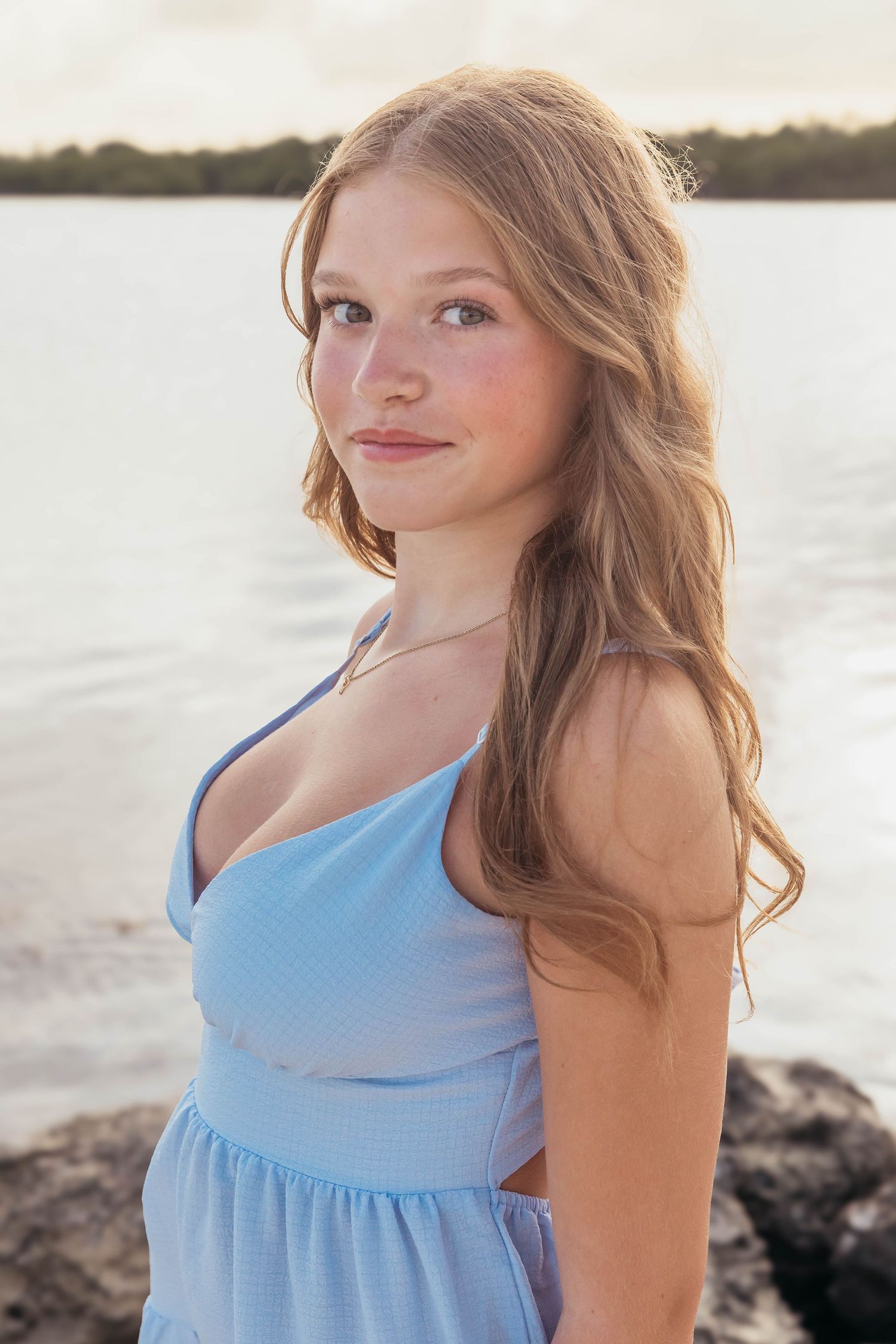 Young woman with long auburn hair wearing a blue sundress poses by a serene waterfront at golden hour.