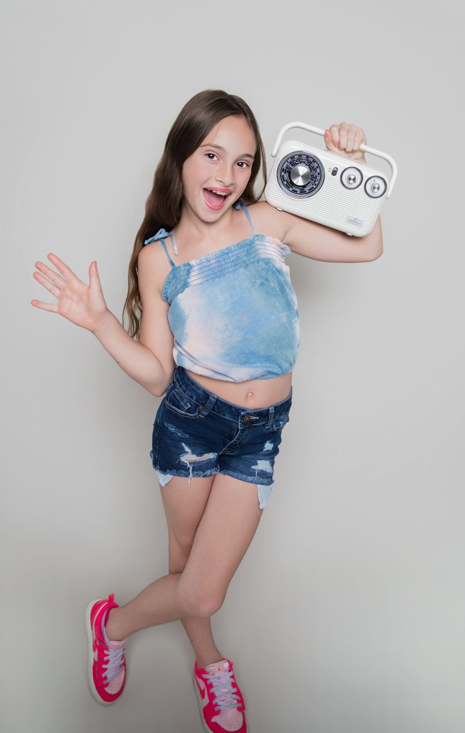 Happy young girl in denim shorts and blue top holding a vintage radio, posing energetically against a gray background.