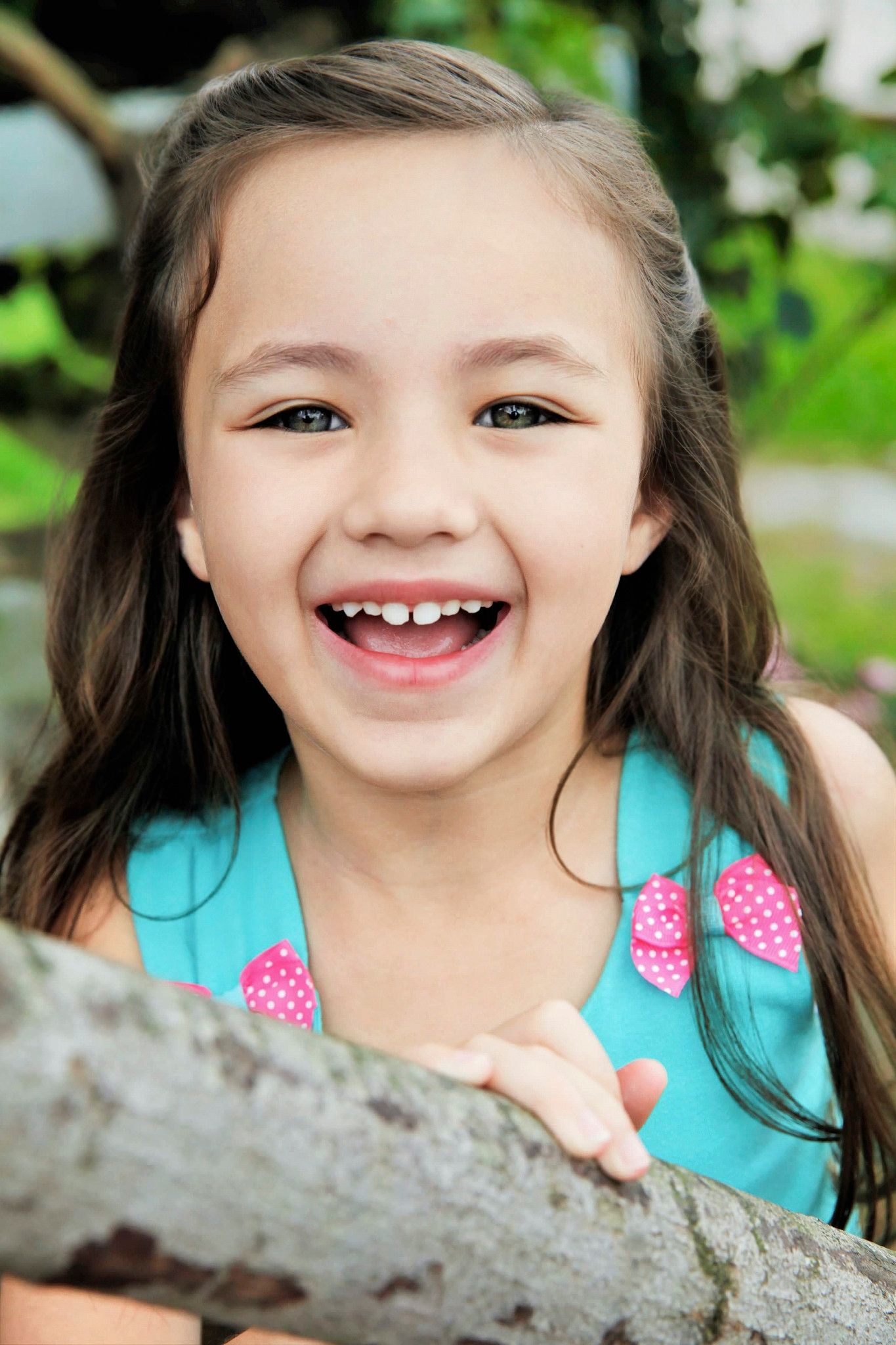 A happy young girl with long dark hair smiles broadly, showing missing teeth, wearing a teal dress outdoors.