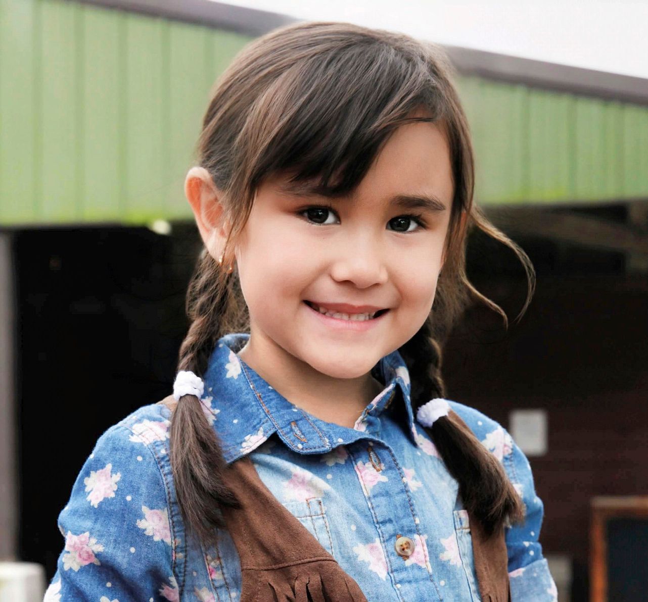 Young girl with braided pigtails smiling outdoors, wearing a blue floral denim shirt.