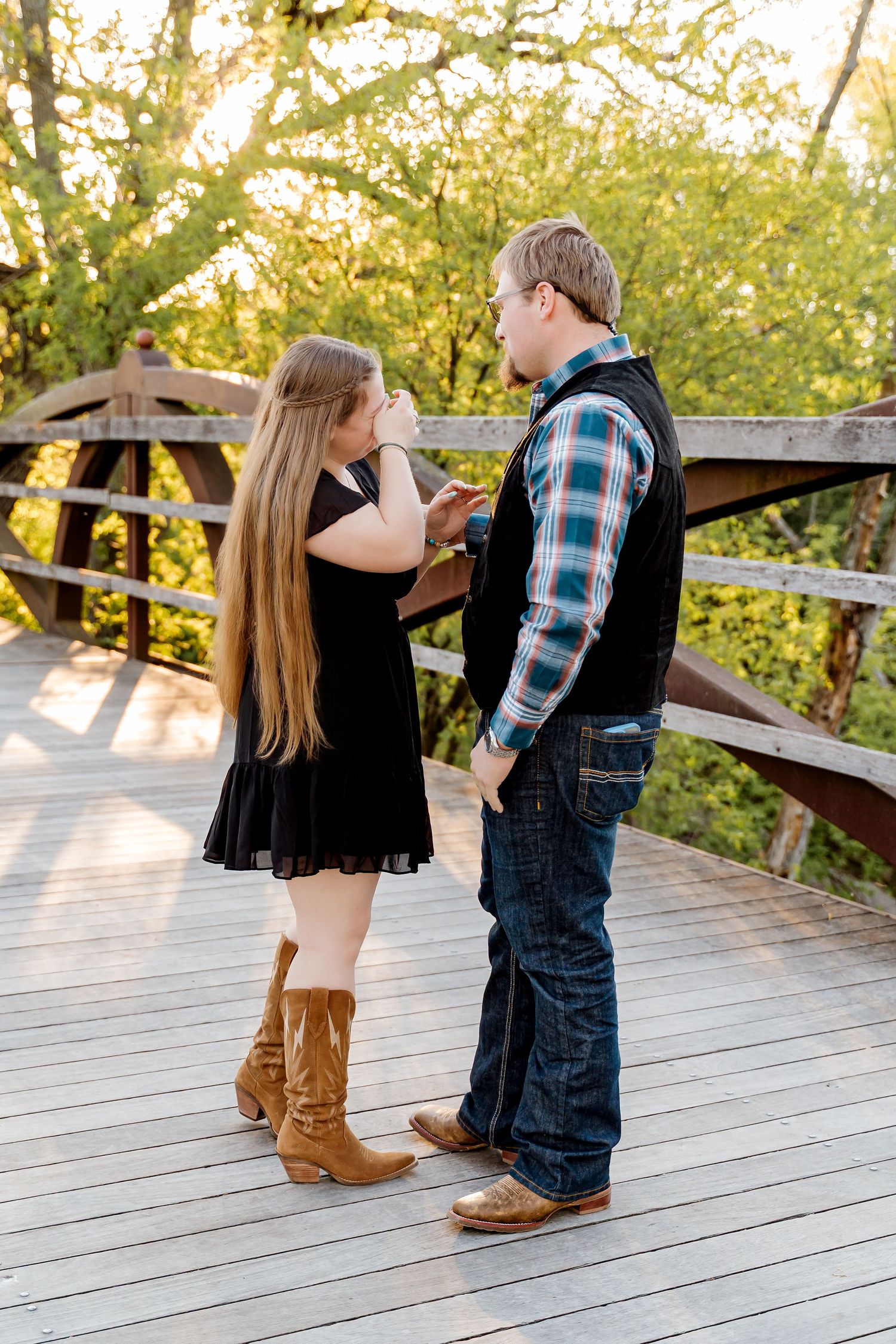 Couple standing on a wooden bridge surrounded by green trees, woman in black dress and cowboy boots facing man in plaid shirt.