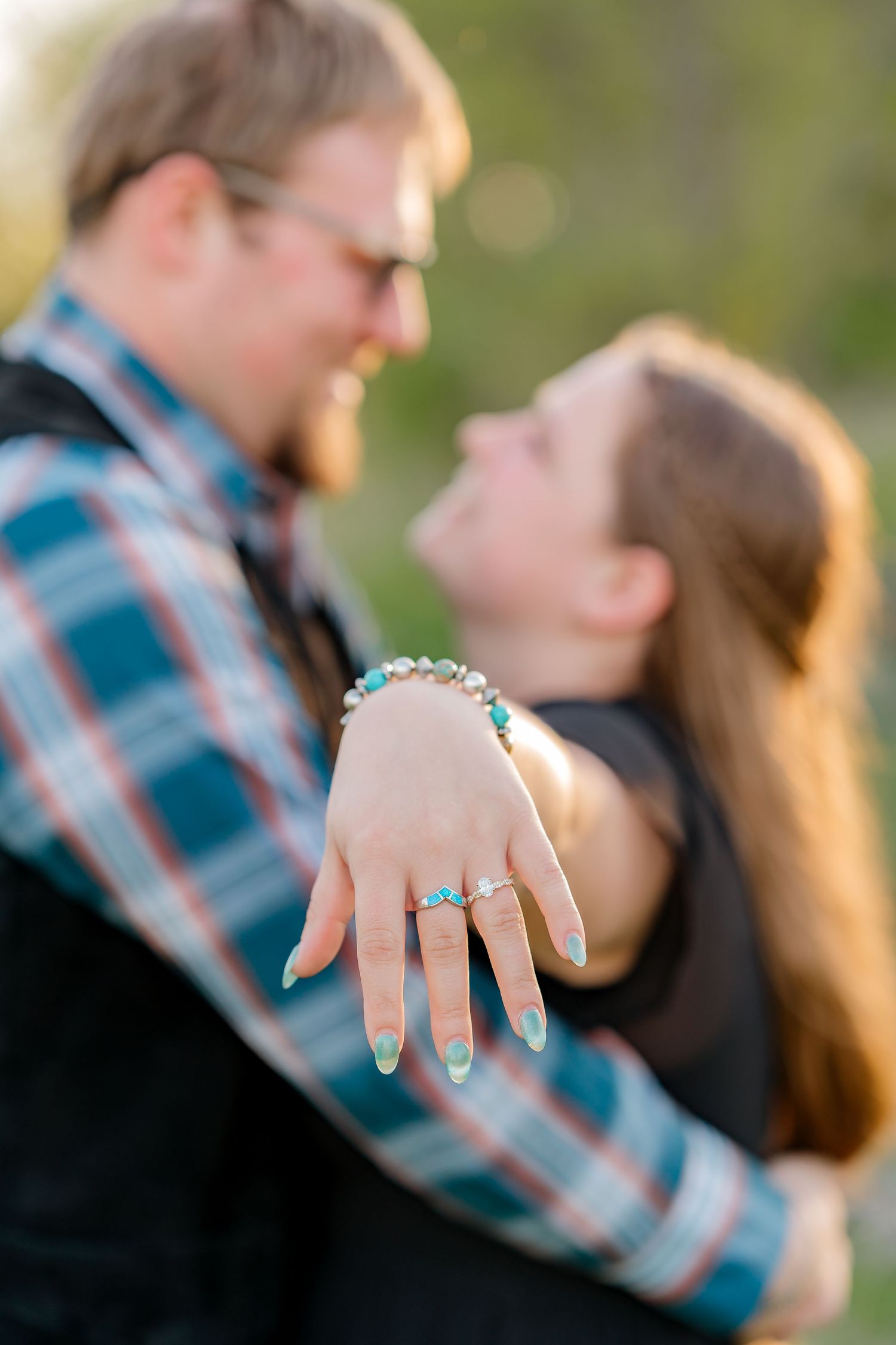 Couple sharing a joyful engagement moment, woman displaying diamond ring with teal nails and bracelet outdoors.