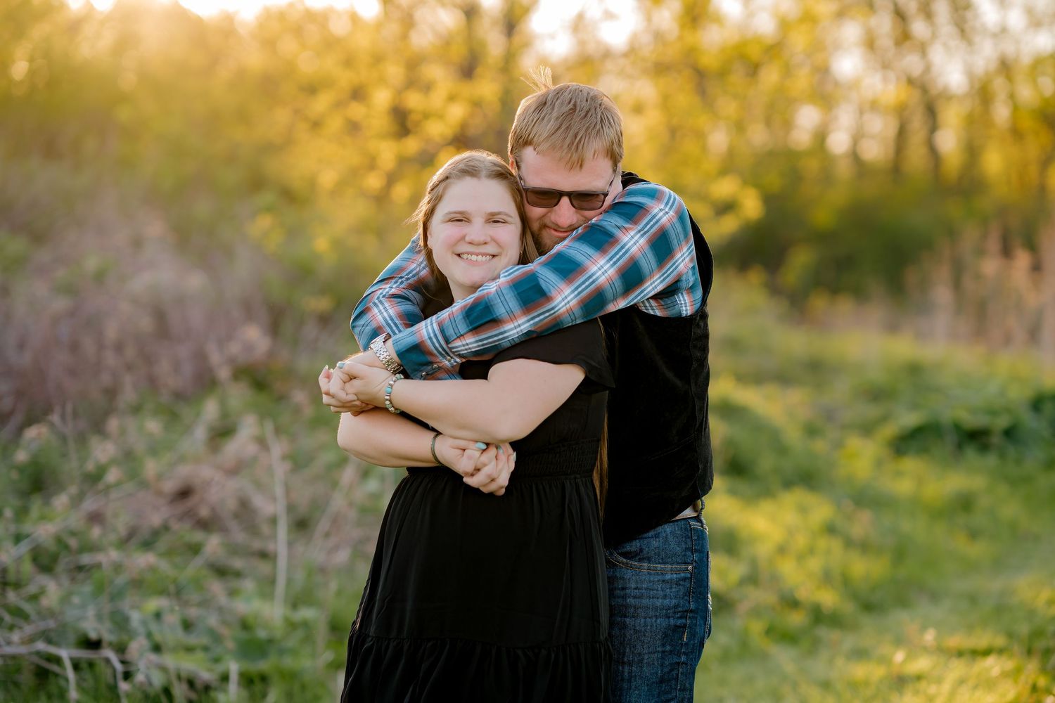 Couple embracing outdoors in golden hour light, man hugging woman from behind in a green field.