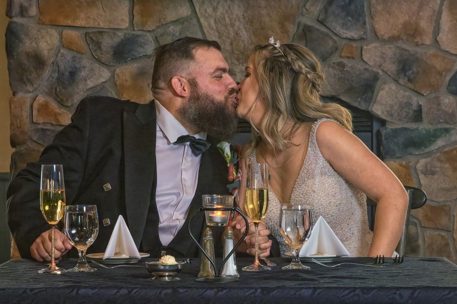 Newlywed couple sharing a kiss at their wedding reception table with champagne glasses and stone wall backdrop.