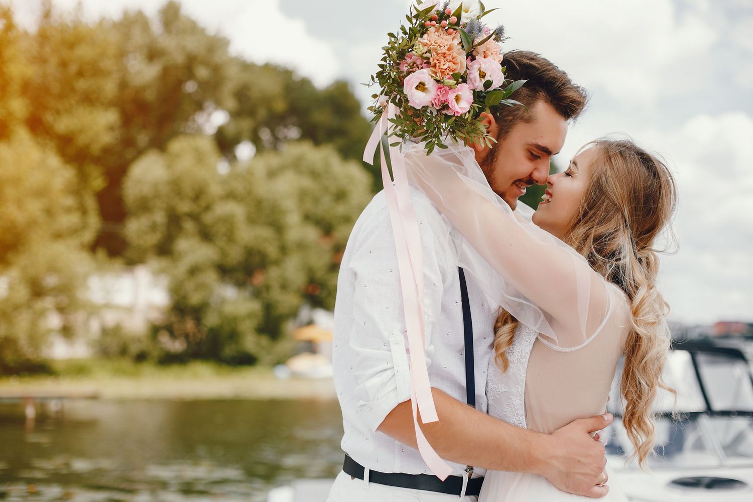 A romantic couple nearly kissing outdoors near a lake, the bride holding a pink floral bouquet.
