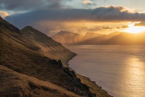 Golden sunset light breaks through storm clouds over a rugged coastal hillside and calm fjord in the Faroe Islands.