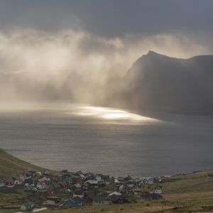 Misty morning light filters through clouds over a colorful Faroese village nestled beside a tranquil mountain fjord.