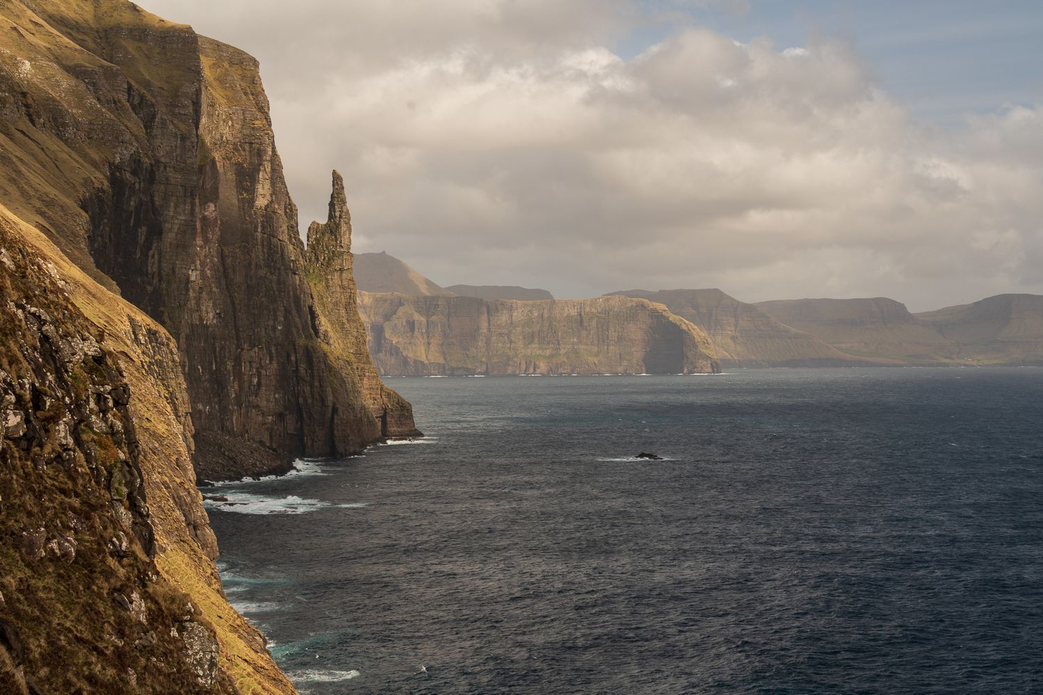 Dramatic sea cliffs with a rock spire rise above the North Atlantic Ocean under cloudy skies in the Faroe Islands.