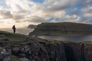 A lone hiker stands on a cliff edge overlooking a dramatic coastal lake and layered rock formations in the Faroe Islands.