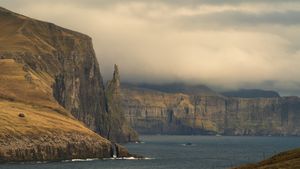 Towering golden-brown sea cliffs stretch along the rugged North Atlantic coastline under moody storm clouds in the Faroe Islands.
