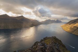 Panoramic view of a winding fjord flanked by steep mountains glowing in warm sunset light in the Faroe Islands.