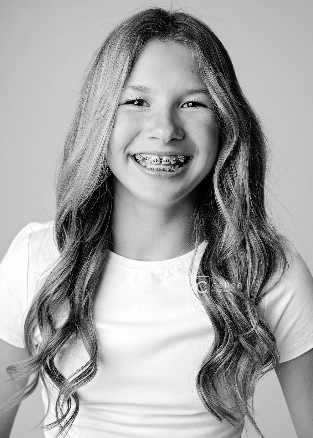 Black and white portrait of a smiling young girl with long wavy hair and braces wearing a white t-shirt.