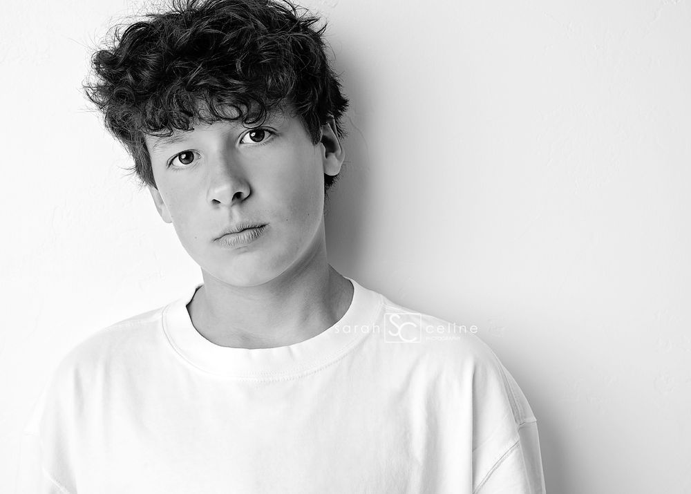 Black and white portrait of a young teenage boy with curly hair wearing a white t-shirt against a light background.