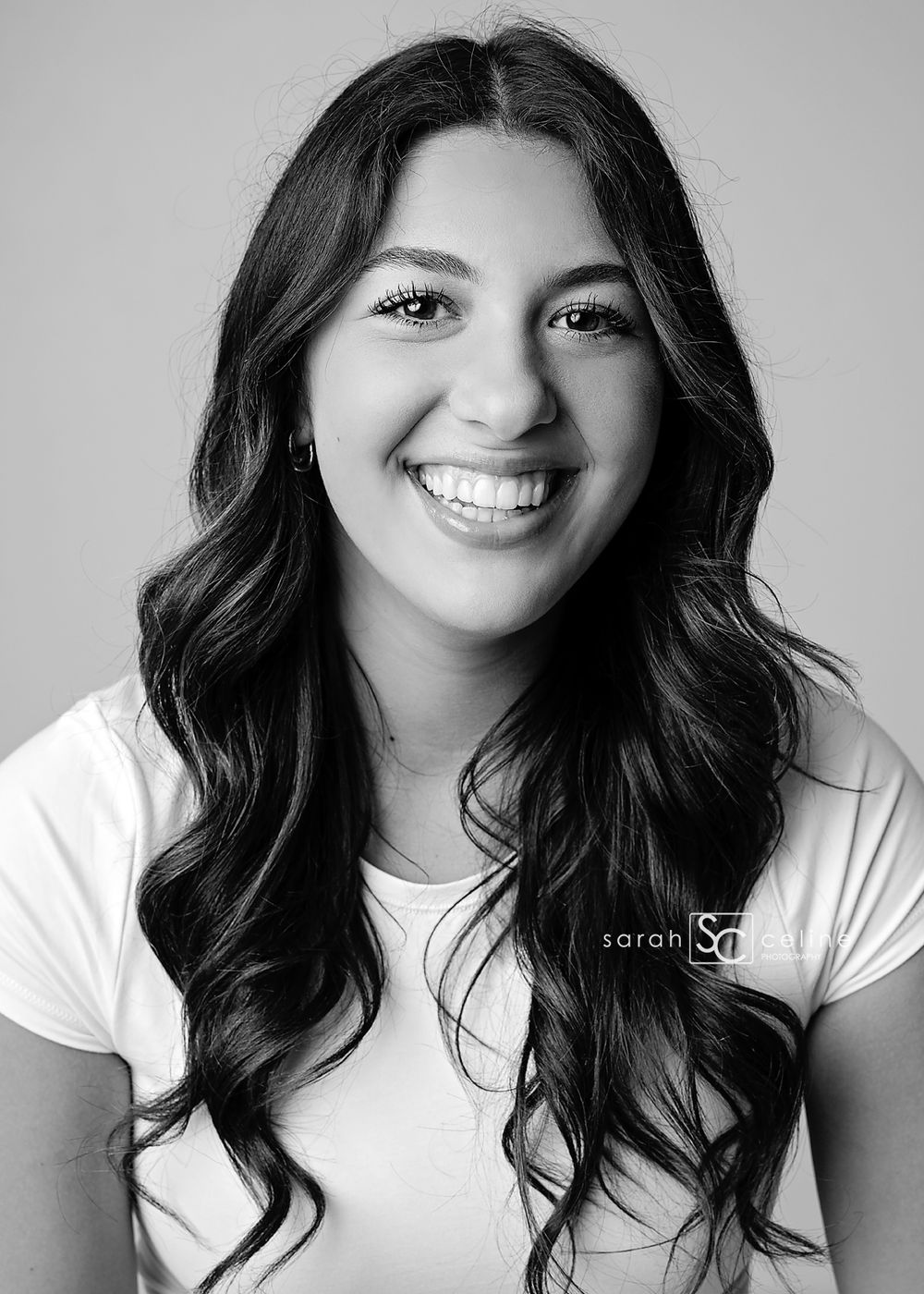 Black and white professional headshot of a young woman with long wavy hair, smiling confidently in a white t-shirt.