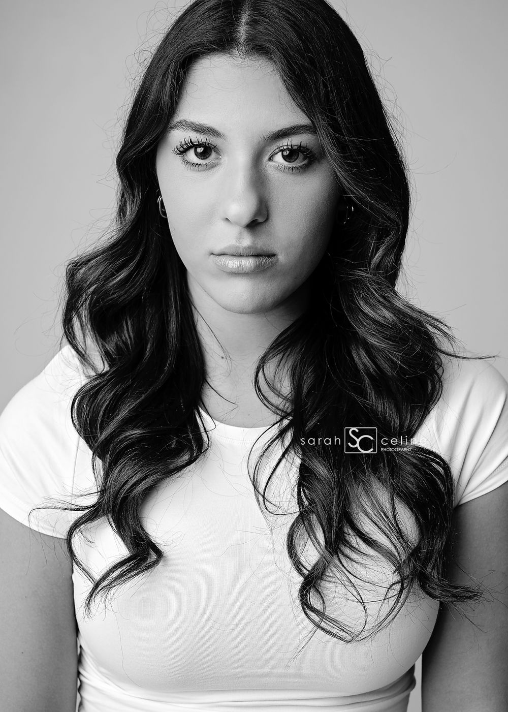 Black and white portrait of a young woman with long wavy dark hair wearing a white top, photographed by Sarah Cell.
