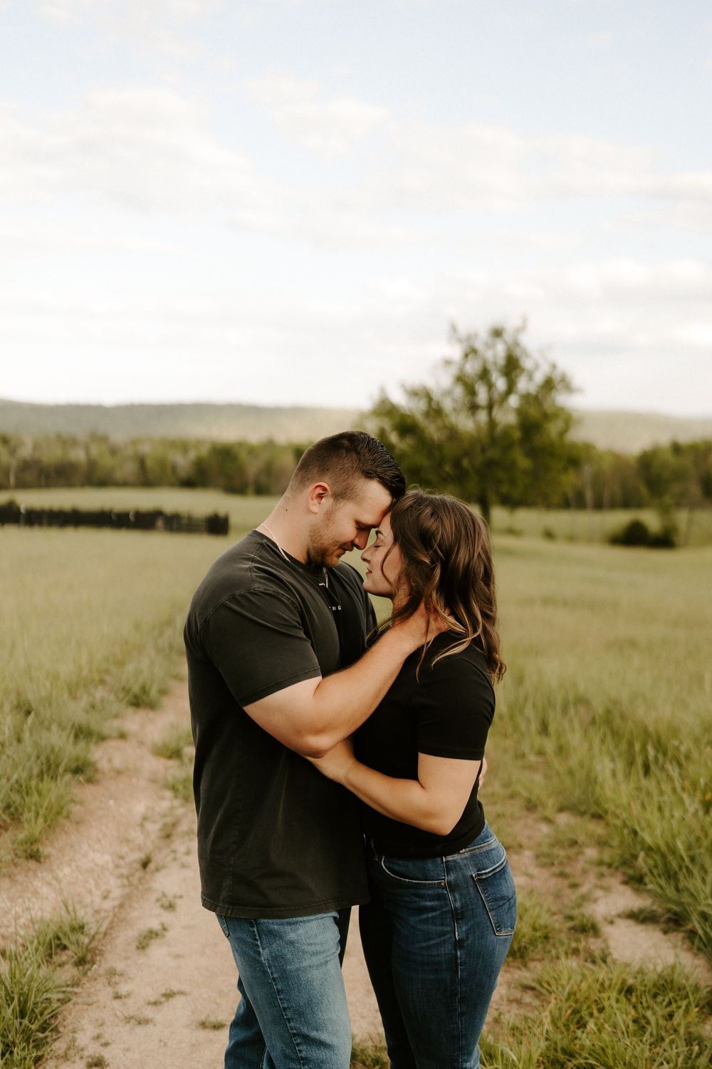 Couple embracing and nearly kissing on a dirt path surrounded by lush green fields at golden hour.