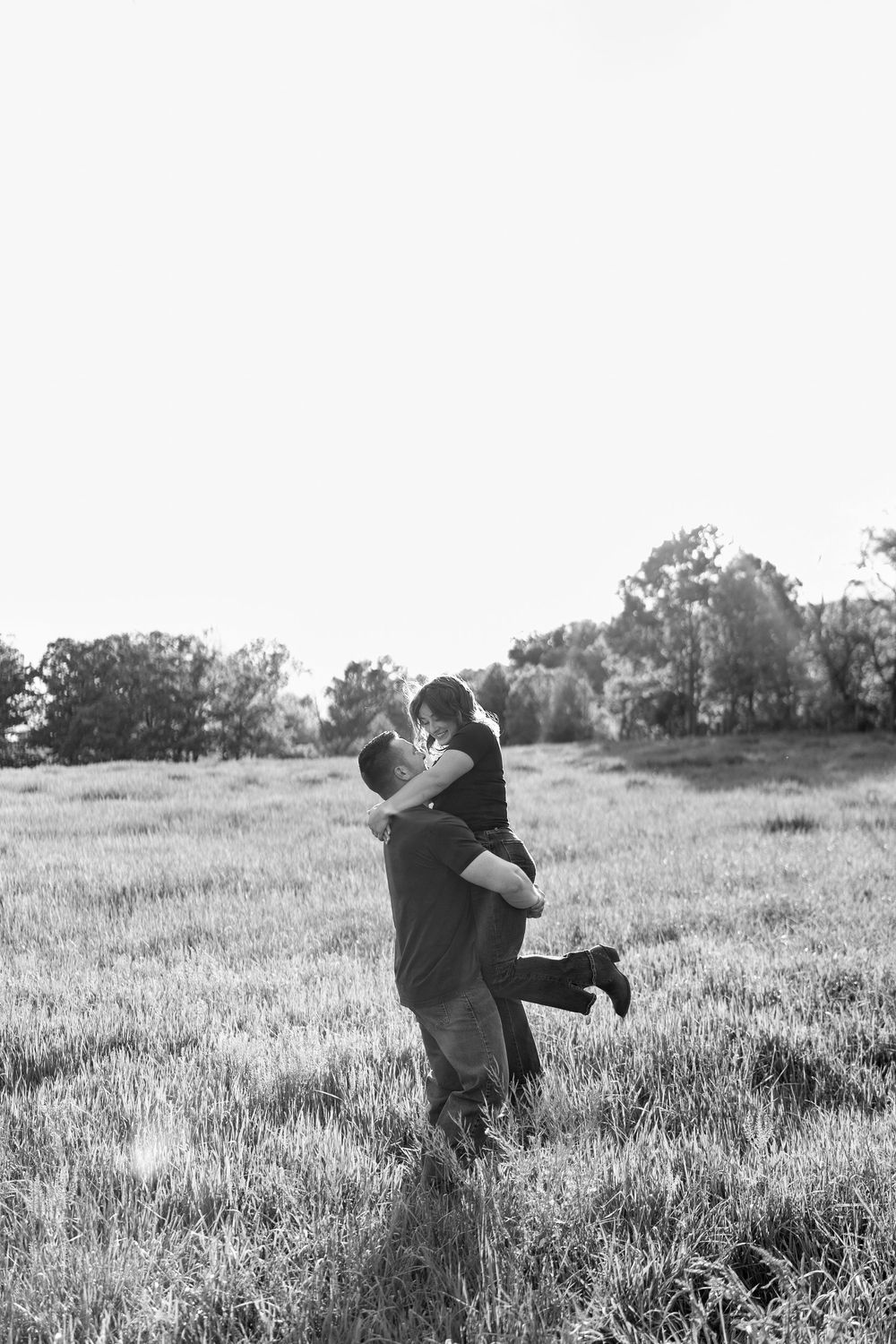 Black and white photo of man lifting woman in arms in a grassy field with trees behind.