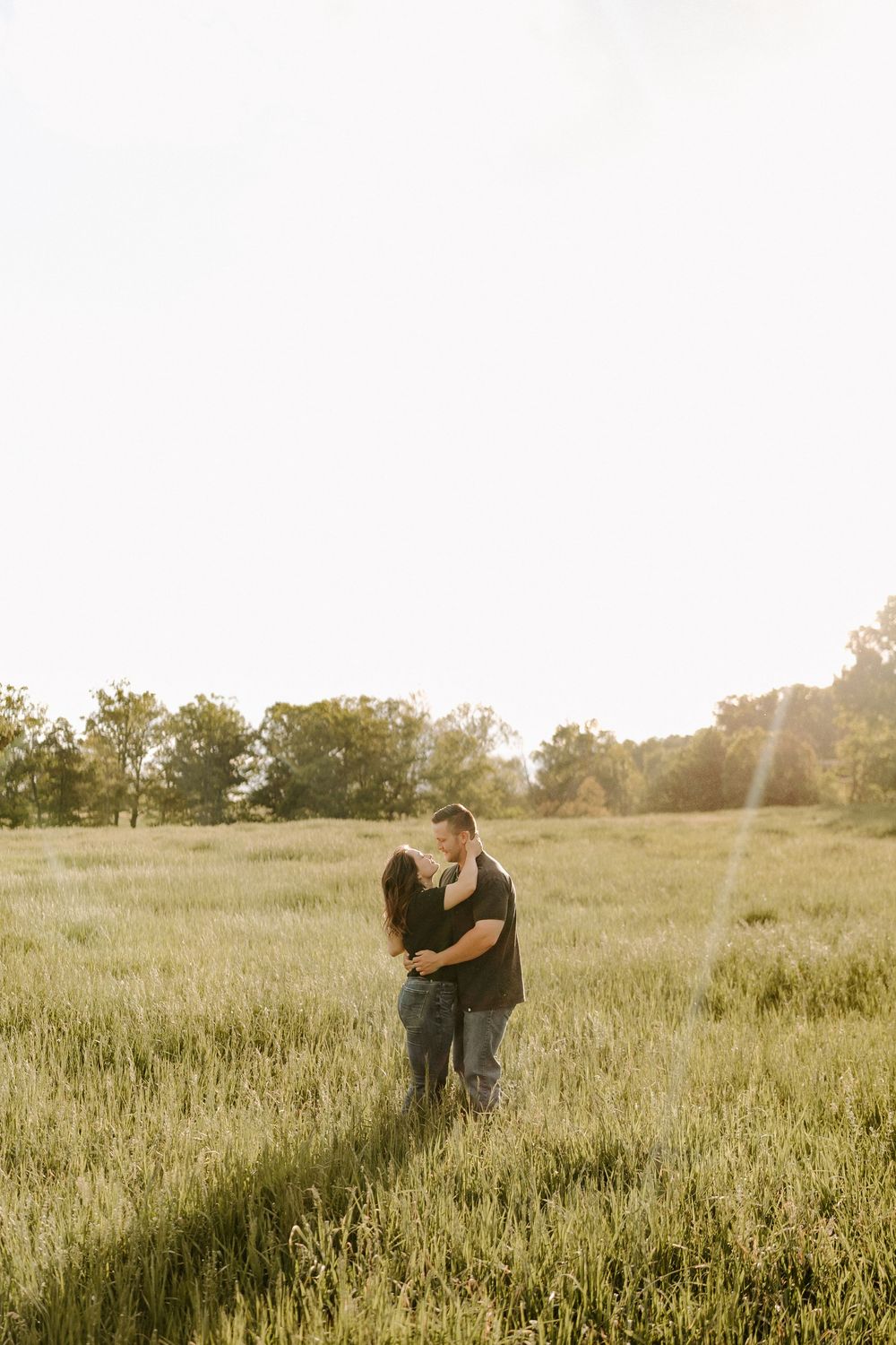 Couple sharing a romantic kiss in a golden sunlit grassy field surrounded by trees.
