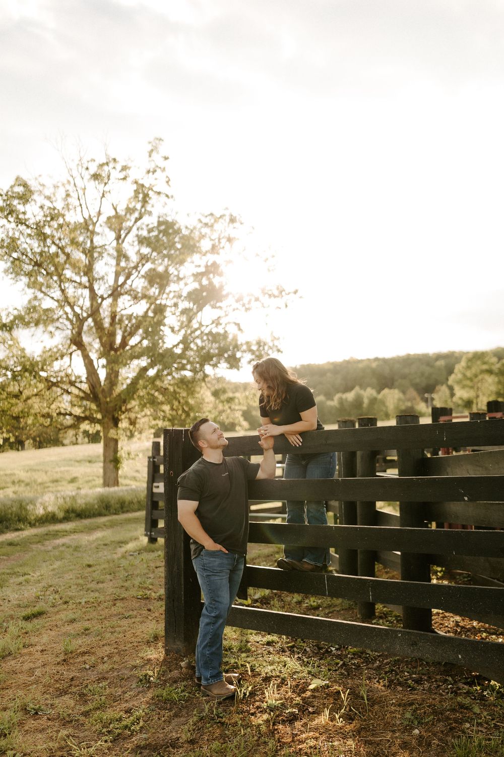 Man gazing up at woman sitting on a rustic wooden fence in a golden sunset field.