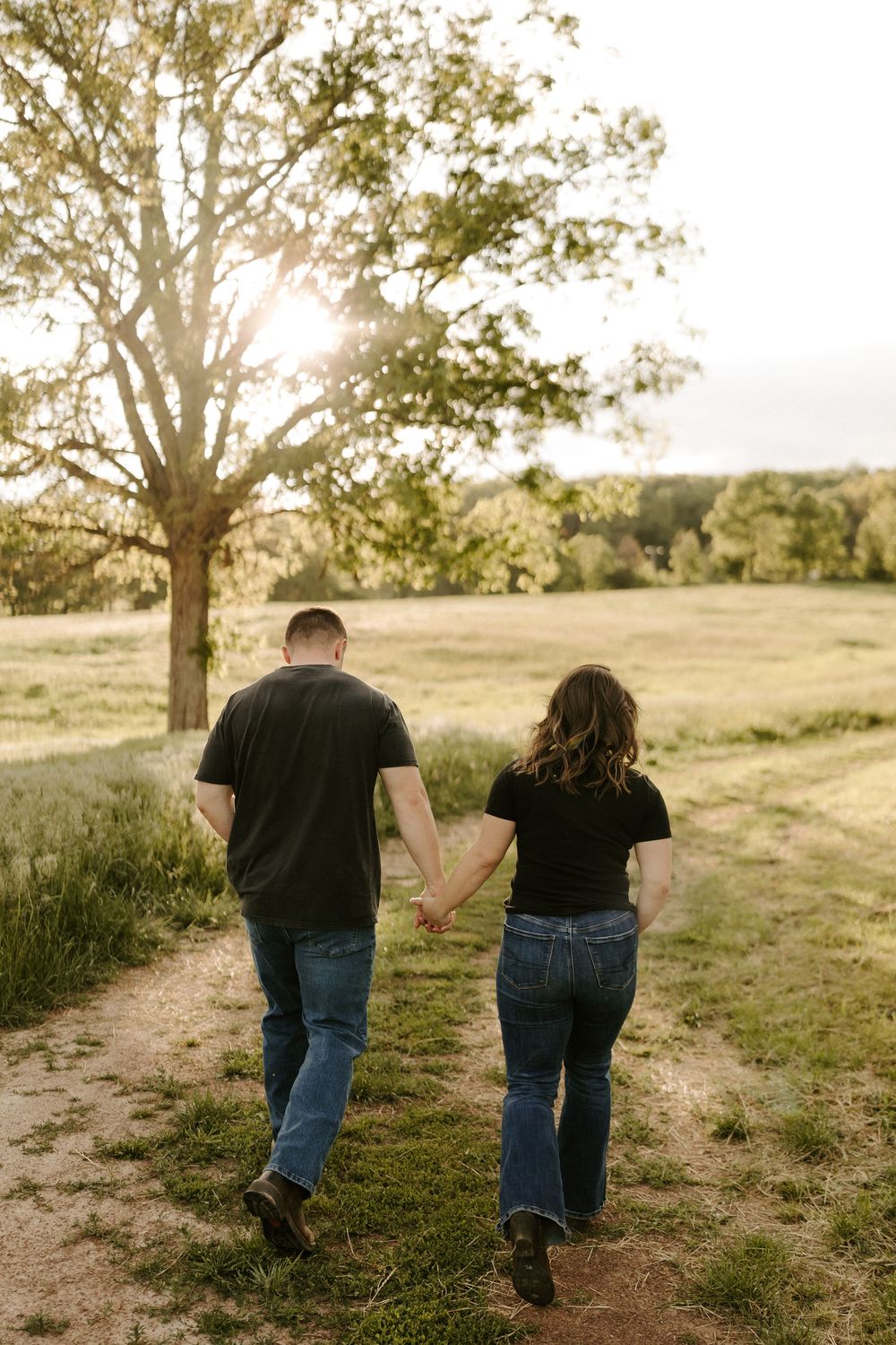 Couple walking hand in hand away from camera on a sunlit path through a scenic green meadow.
