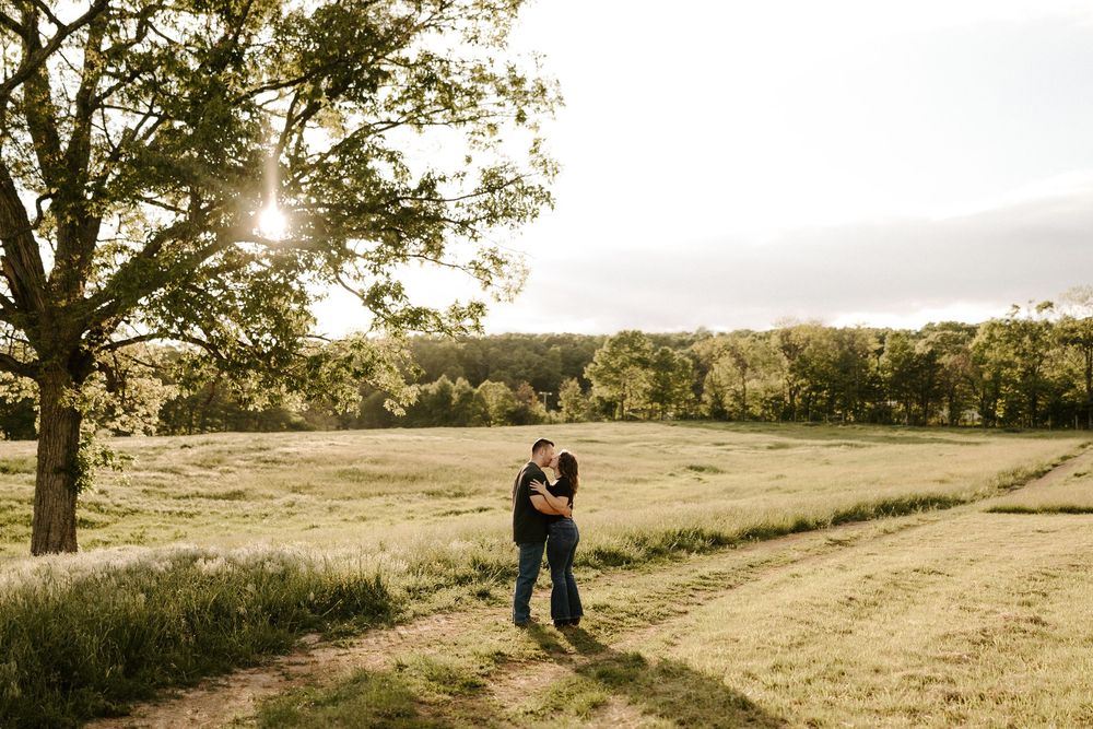 Couple embracing near a large tree in a wide open sunlit farm field at golden hour.