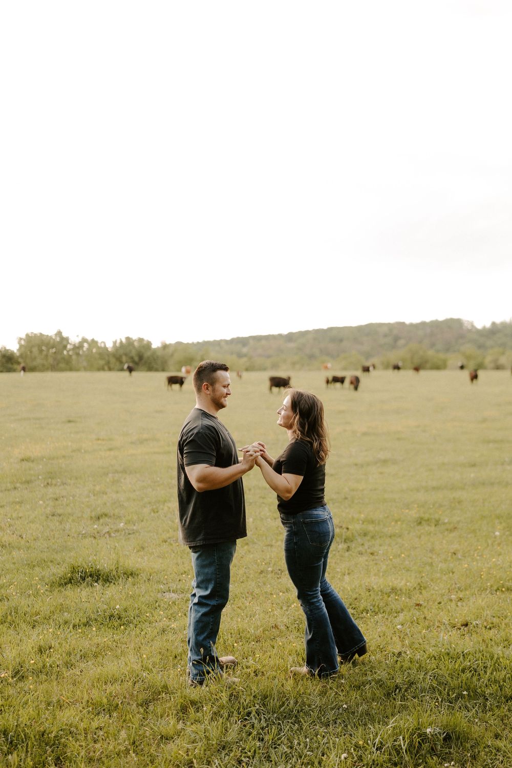 Smiling couple holding hands and dancing in a green pasture with cattle in background.
