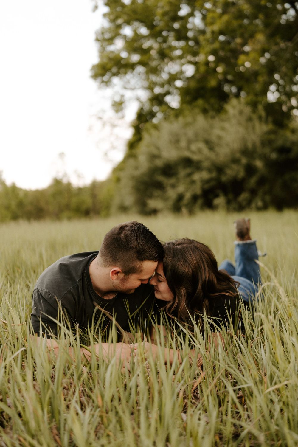 Couple lying in tall green grass touching foreheads romantically during outdoor engagement photo session.