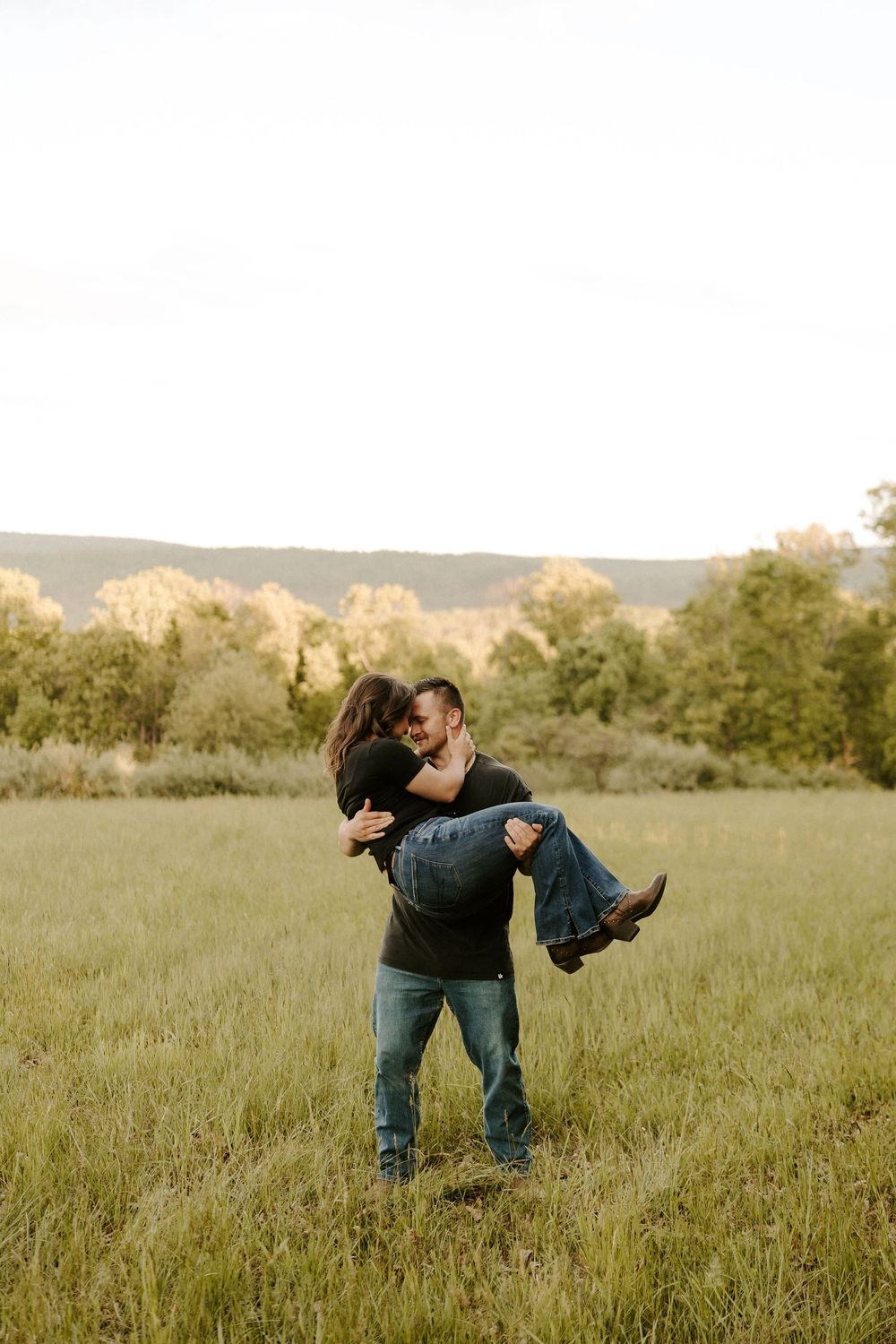 Man lifting woman in his arms in a green meadow during a romantic outdoor engagement photo session.