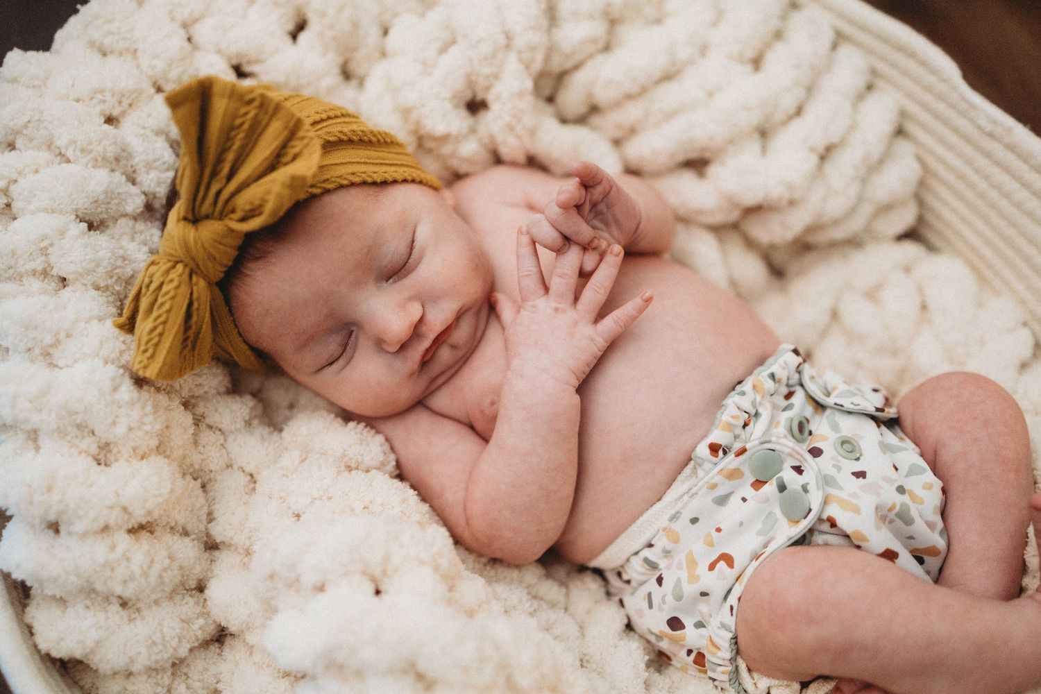 Smiling newborn baby girl sleeping on fluffy white blanket wearing floral shorts and mustard yellow bow headband.