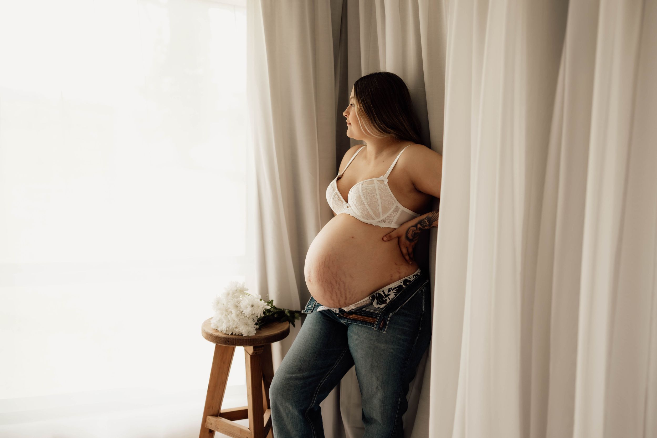 Pregnant woman in white lace bra and jeans standing by sheer curtains, cradling her baby bump near a wooden stool.