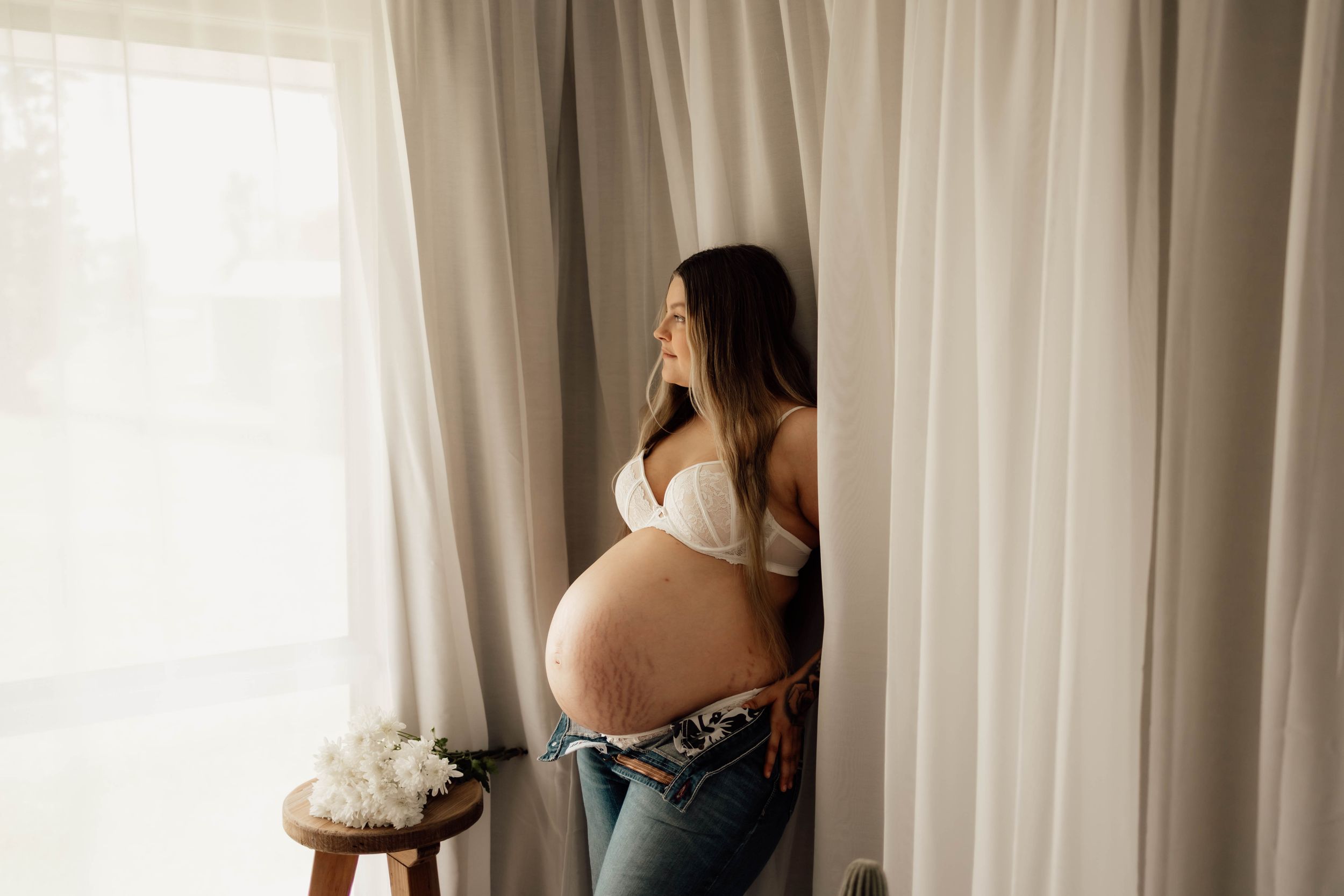 Pregnant woman in white bra and jeans cradling baby bump by sheer curtains in natural light.