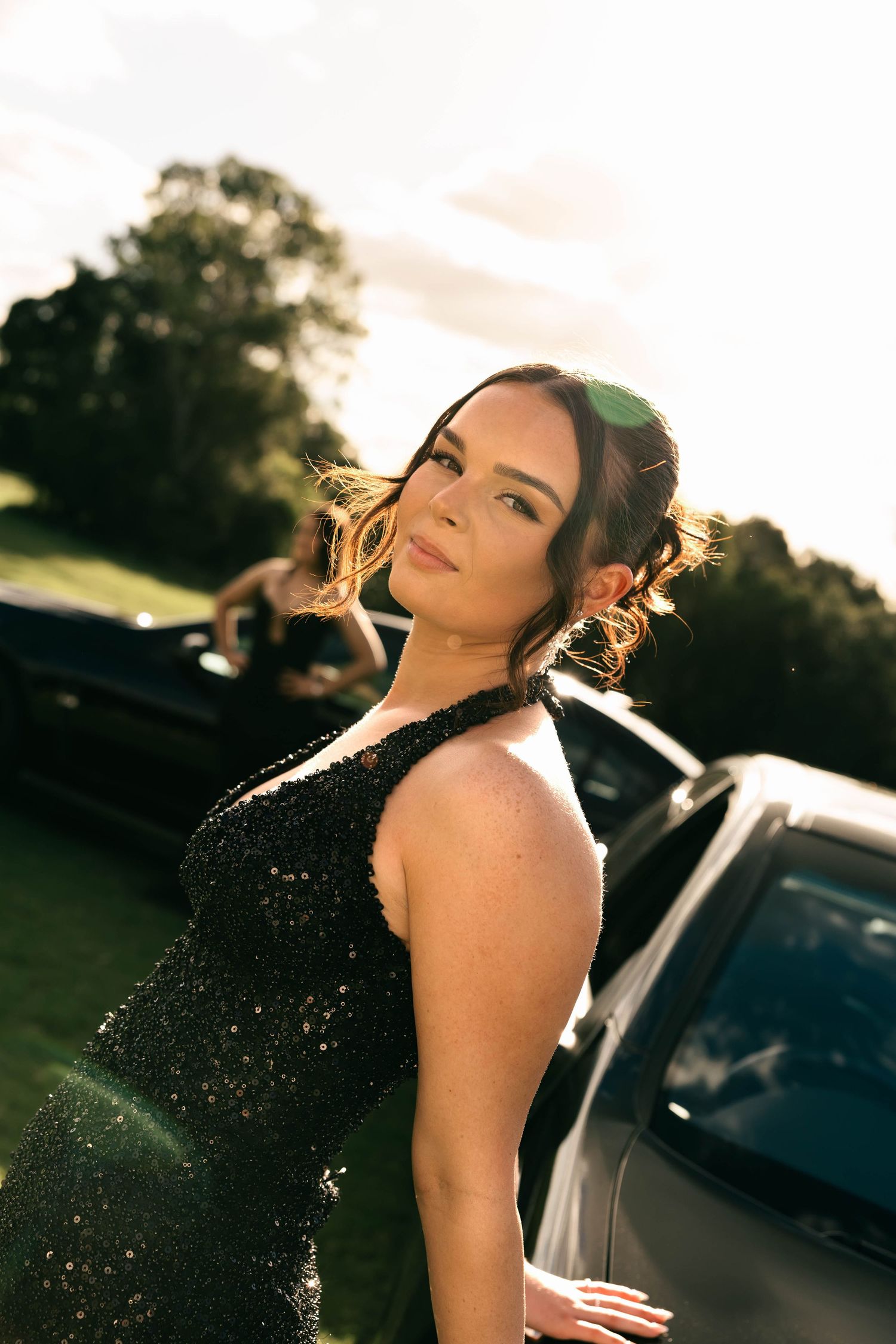 A woman in a black sequin gown poses elegantly near a car outdoors, with golden sunlight illuminating her updo hairstyle.