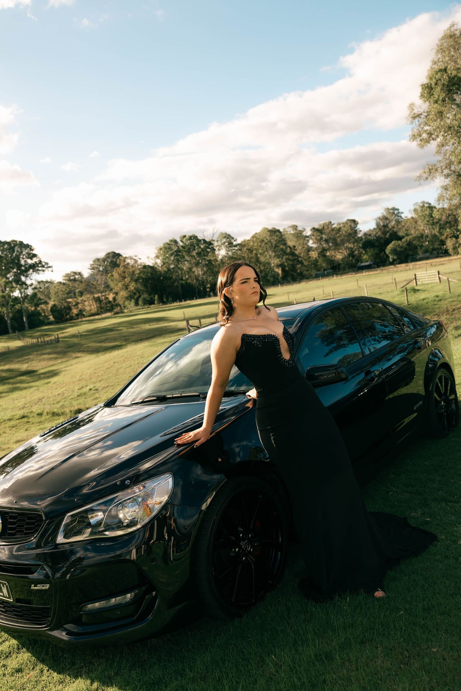Woman in black gown posing beside a black Holden sedan on lush green grass with trees and blue sky.