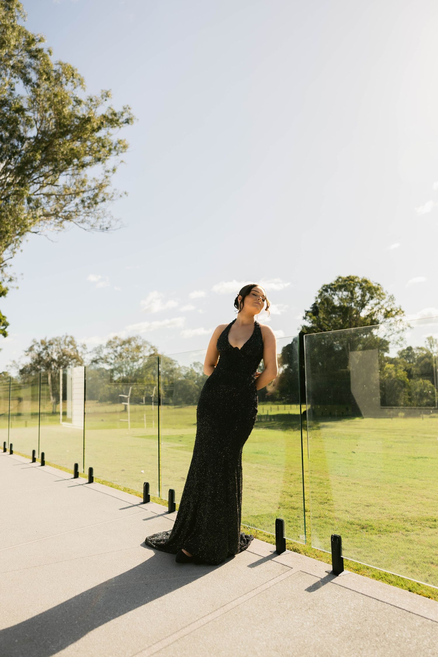 Woman in black lace gown poses by glass fence in sunlit park setting.