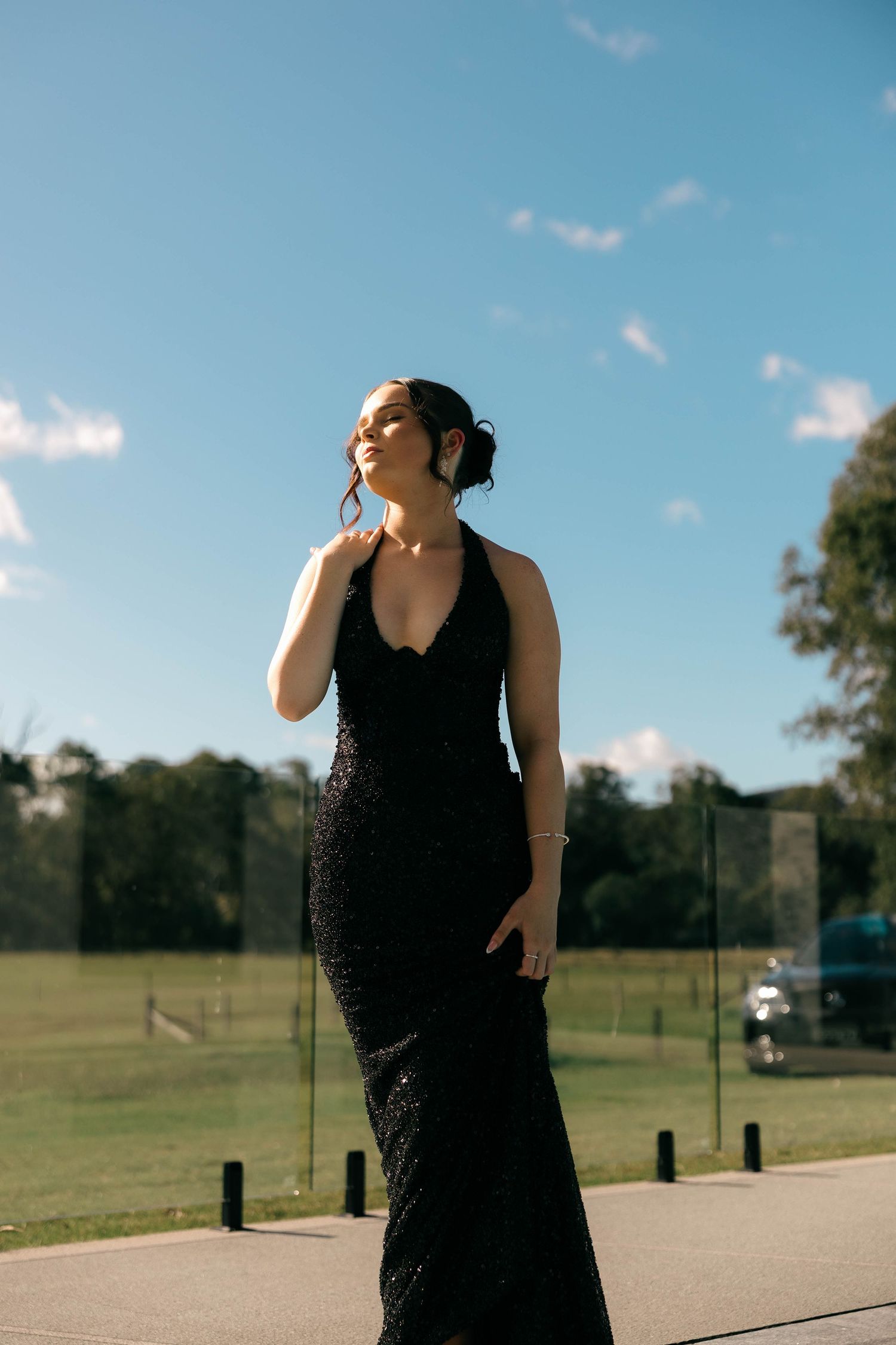 Woman in black sequin halter gown poses outdoors under blue sky on sunny day.