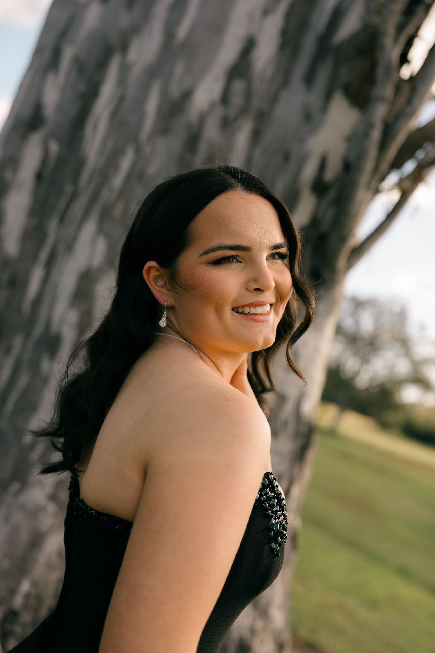 Young woman in black strapless beaded gown smiling outdoors near tree at golden hour.