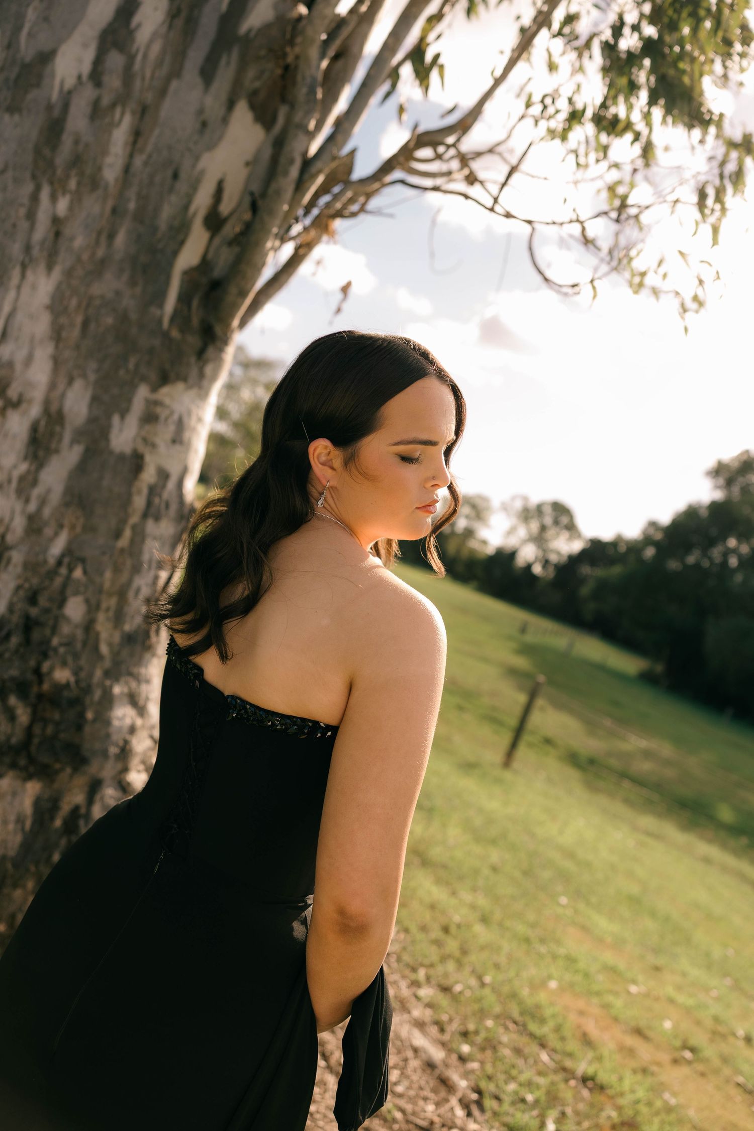 Young woman in black strapless sequin gown leaning against eucalyptus tree in sunlit green field.