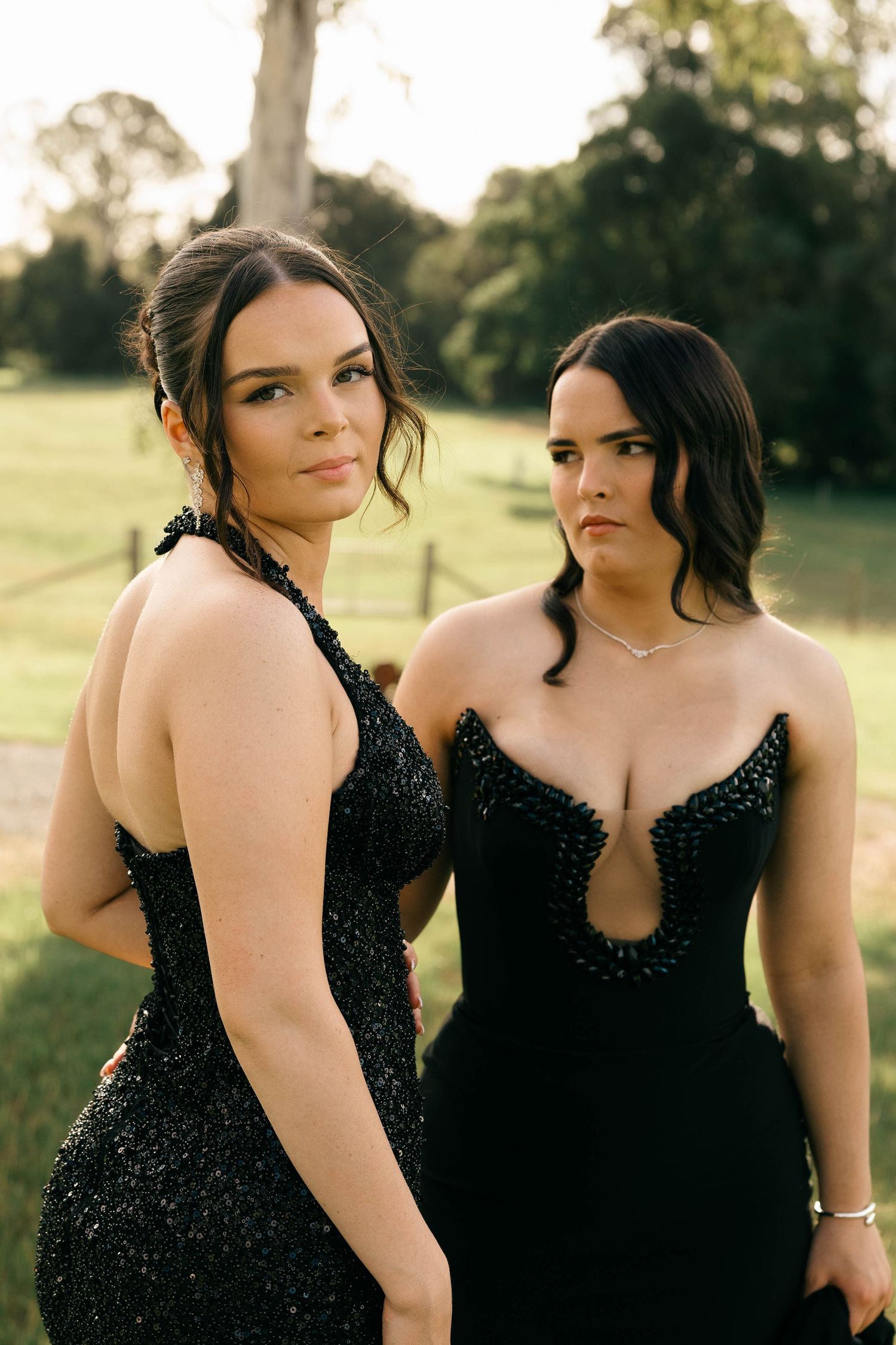 Two women in elegant black beaded formal gowns posing outdoors at golden hour.