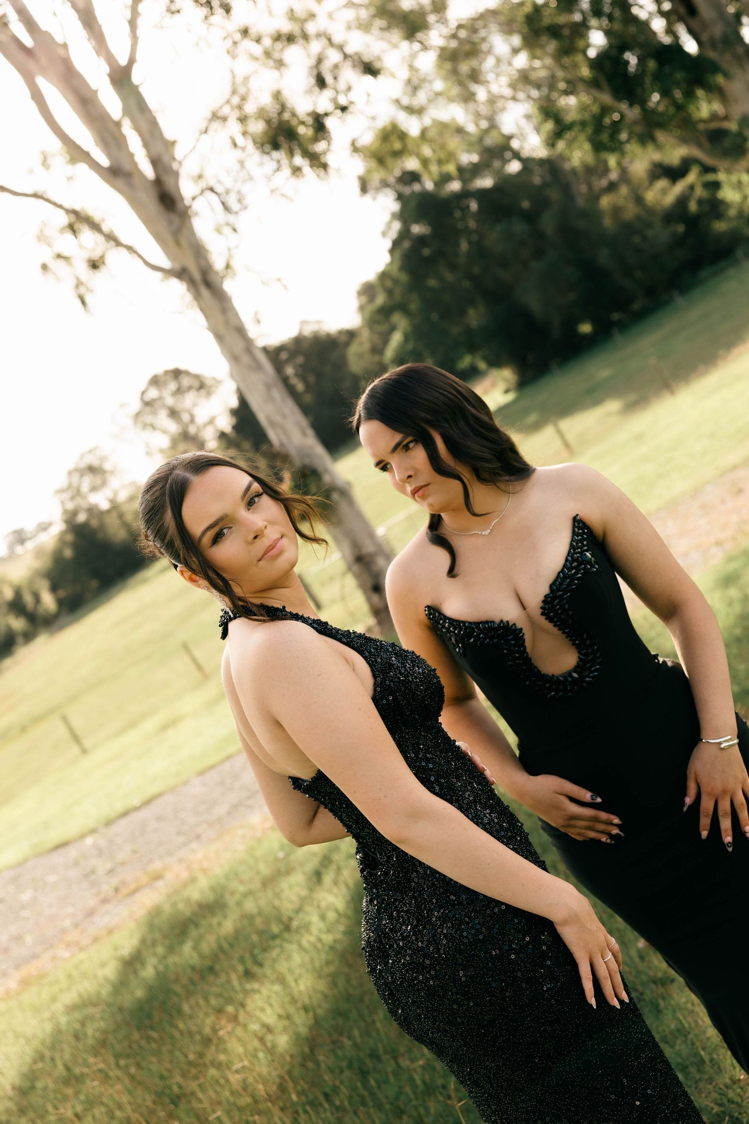 Two women in elegant black sequin gowns posing together in a sunlit outdoor garden setting.