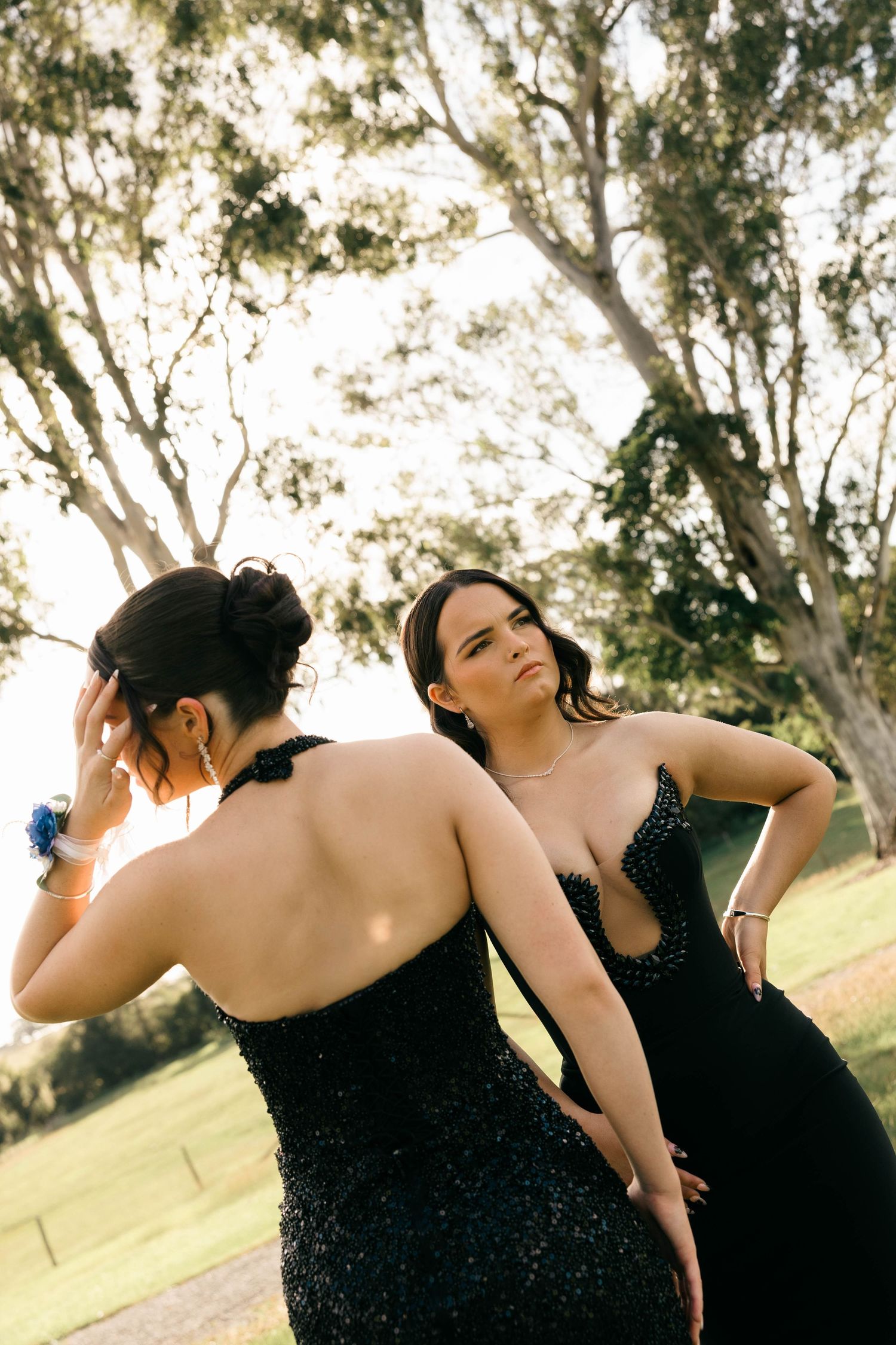 Two women in black beaded formal gowns posing back to back in a park setting.