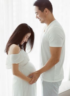 A smiling couple gently cradling a pregnant belly while standing by a bright window.