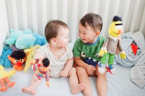 Two baby boys sitting on a white bed surrounded by colorful stuffed toys, smiling at each other.