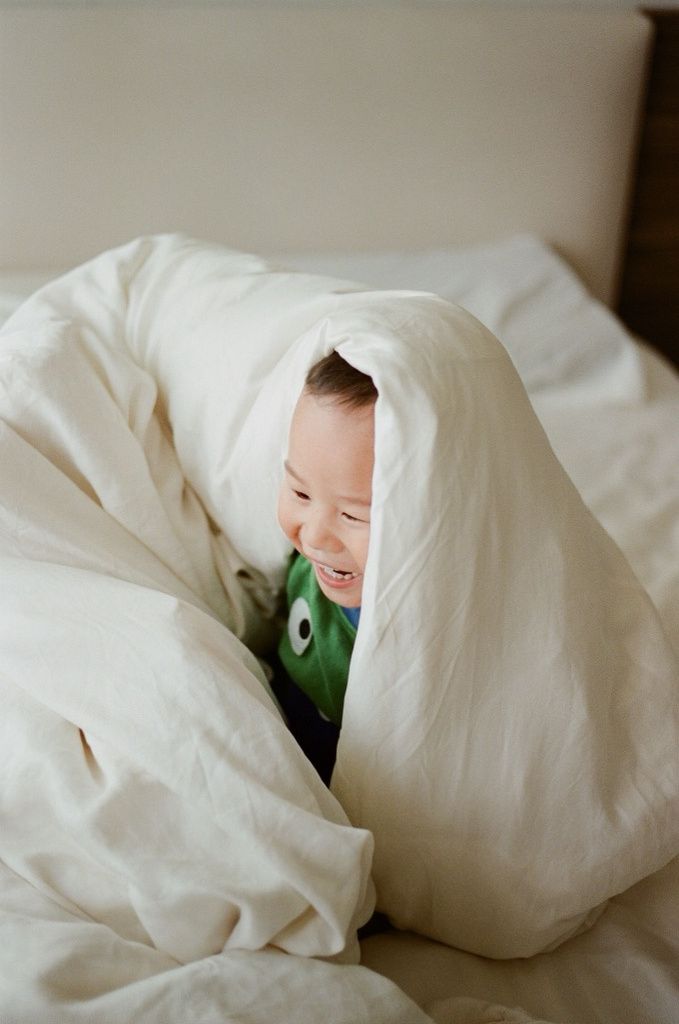 Laughing baby peeking out from fluffy white bedding on a cozy bed.