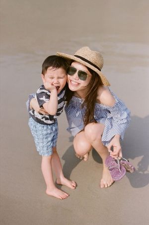 Happy mother crouching on beach with laughing toddler son wearing straw hat.
