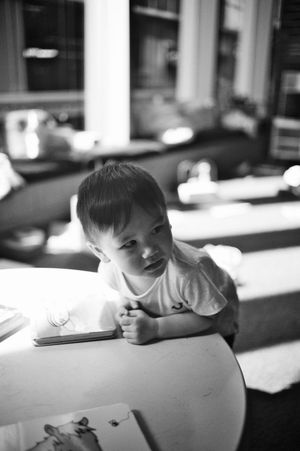 Young boy sitting at a white table, looking down thoughtfully in a black and white photo.