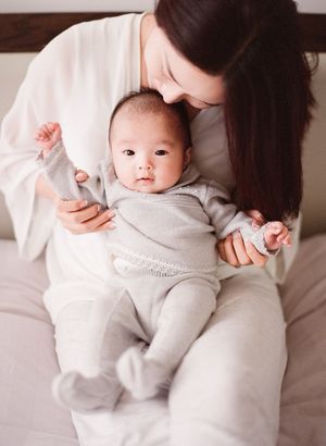 Mother in white holding her alert newborn baby dressed in a gray knit outfit on a bed.